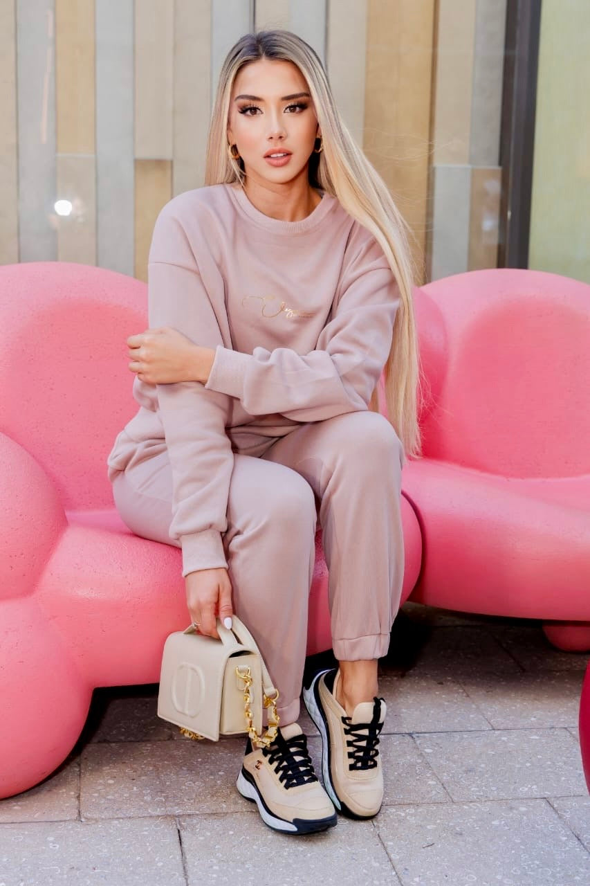 Chic woman in khaki 3-piece jogger set sitting on pink couch; FLY IN STYLE casual outfit.