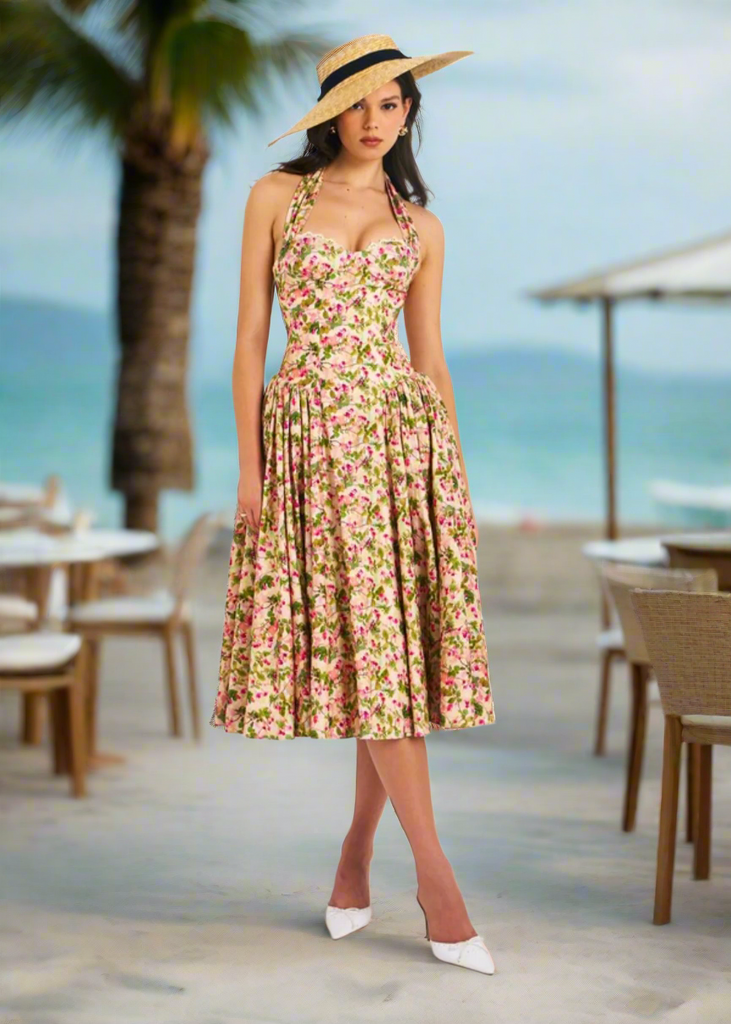 A woman wearing a floral corset midi dress with a pleated skirt, halter neckline, and tie-back detail, paired with a wide-brimmed hat and white shoes.
