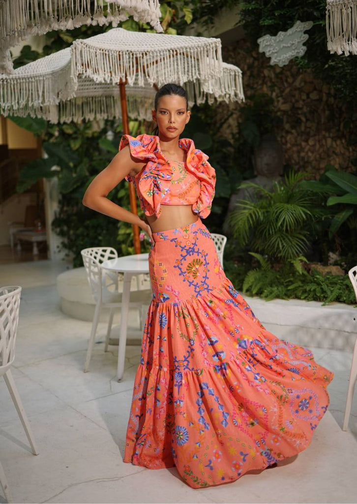 A woman wearing an orange ruffle top and matching maxi skirt with a Côte d’Azur print, standing in a garden setting.