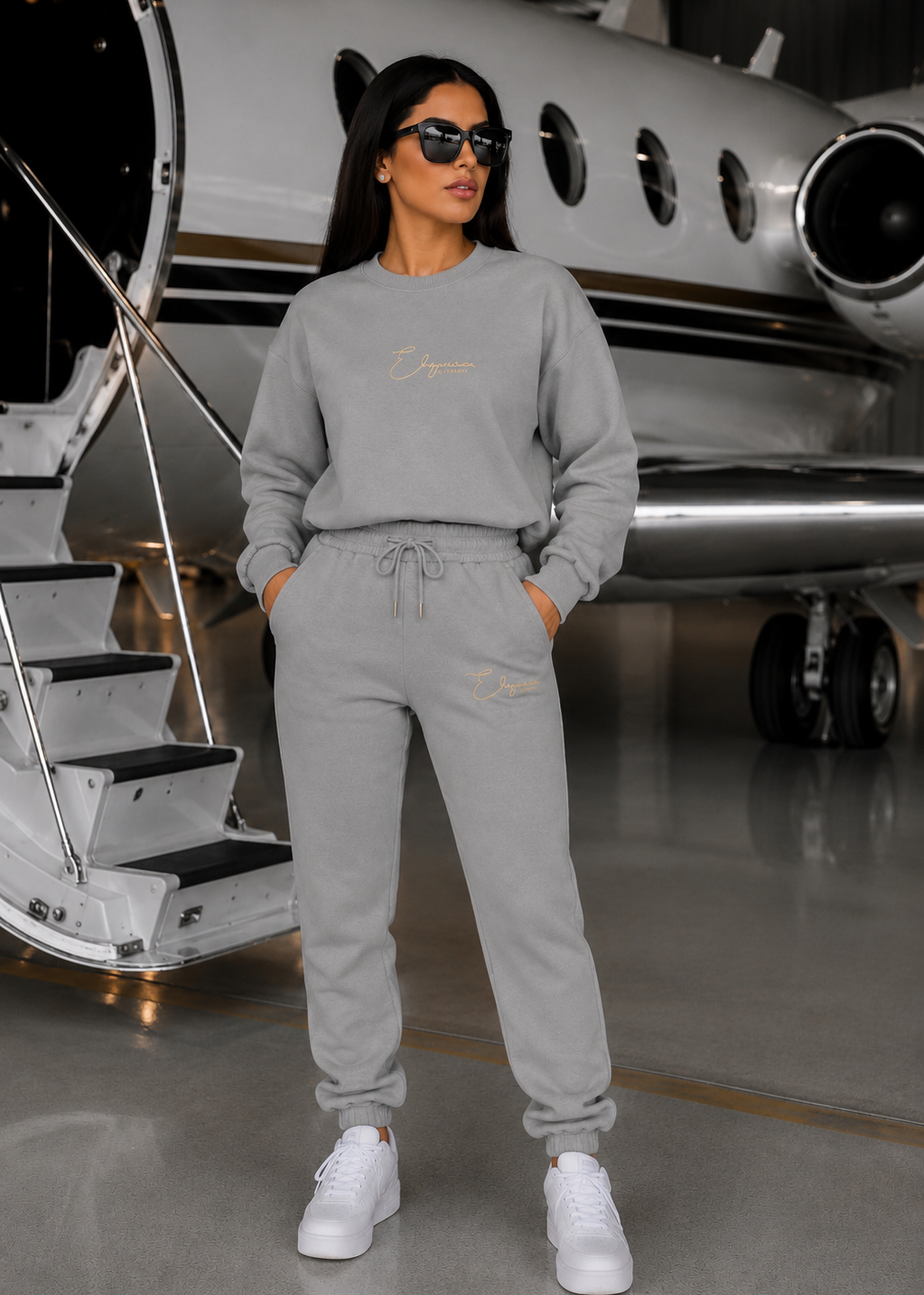 Woman wearing the SNUG SQUAD Grey 3-Piece Travel Jogger Set featuring a luxury grey sweatshirt with gold logo, matching joggers and white sneakers in a private jet hangar.